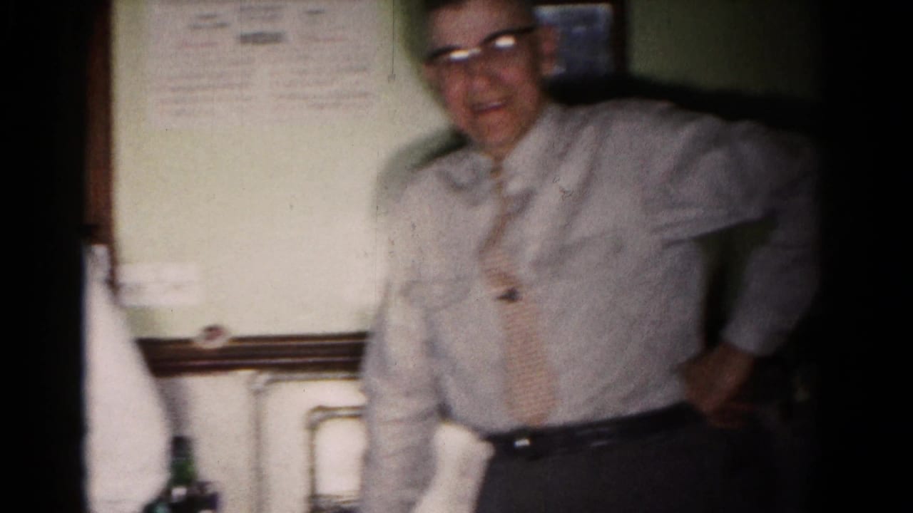 Man in glasses standing in a dim room beside a wall sign and bottles.