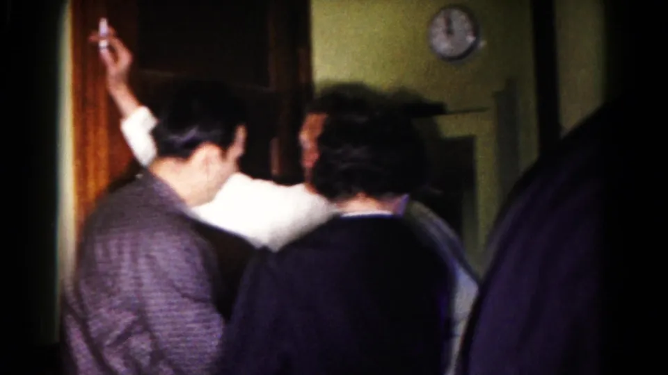 1958 Hagerstown home-movie frame showing adults turned together near curtains, one raising a cigarette in a dim room.