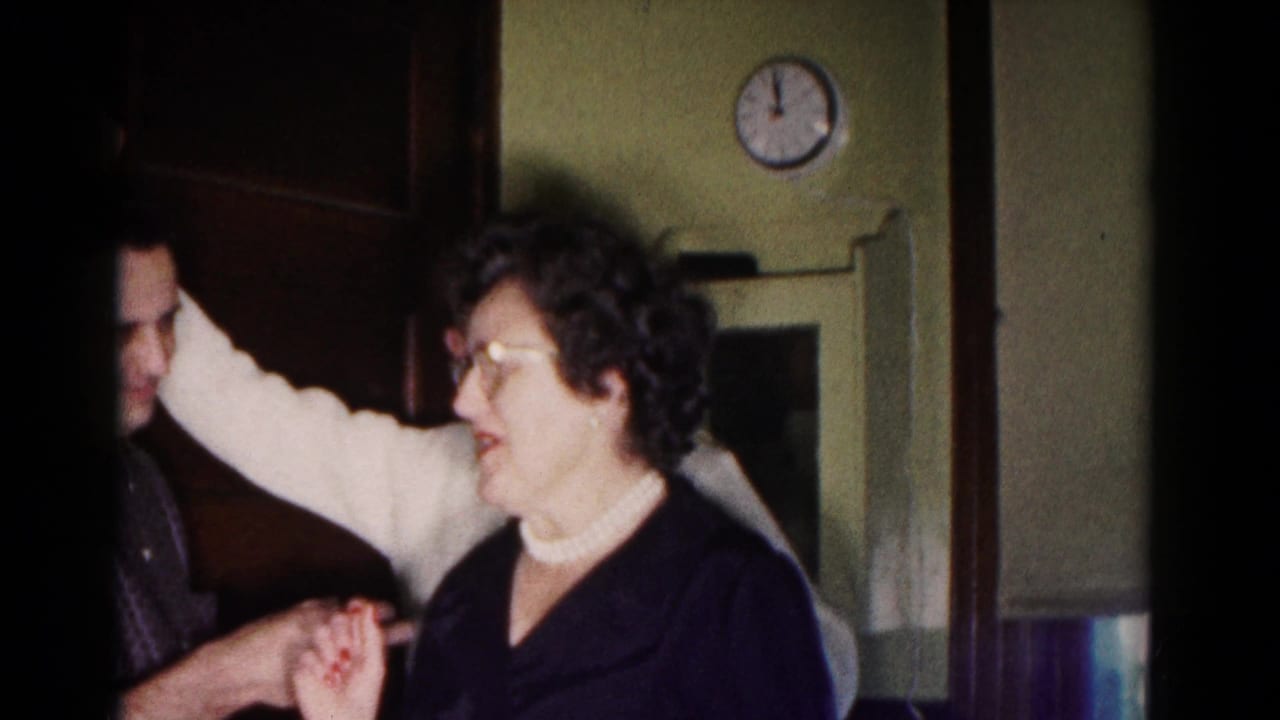 Woman in glasses and pearls laughs indoors while people stand close around her.