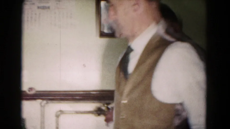 1958 Hagerstown home-movie frame showing a man in a vest walking near a wall calendar in an interior room.
