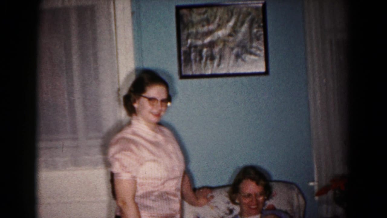 In a 1958 Hagerstown home interior, a woman in glasses stands near a seated woman beneath framed wall art.
