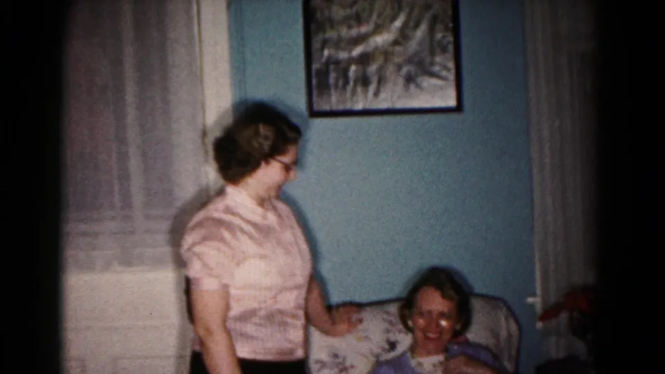 A woman in a striped blouse and glasses turns toward a smiling seated woman in a Hagerstown living room.