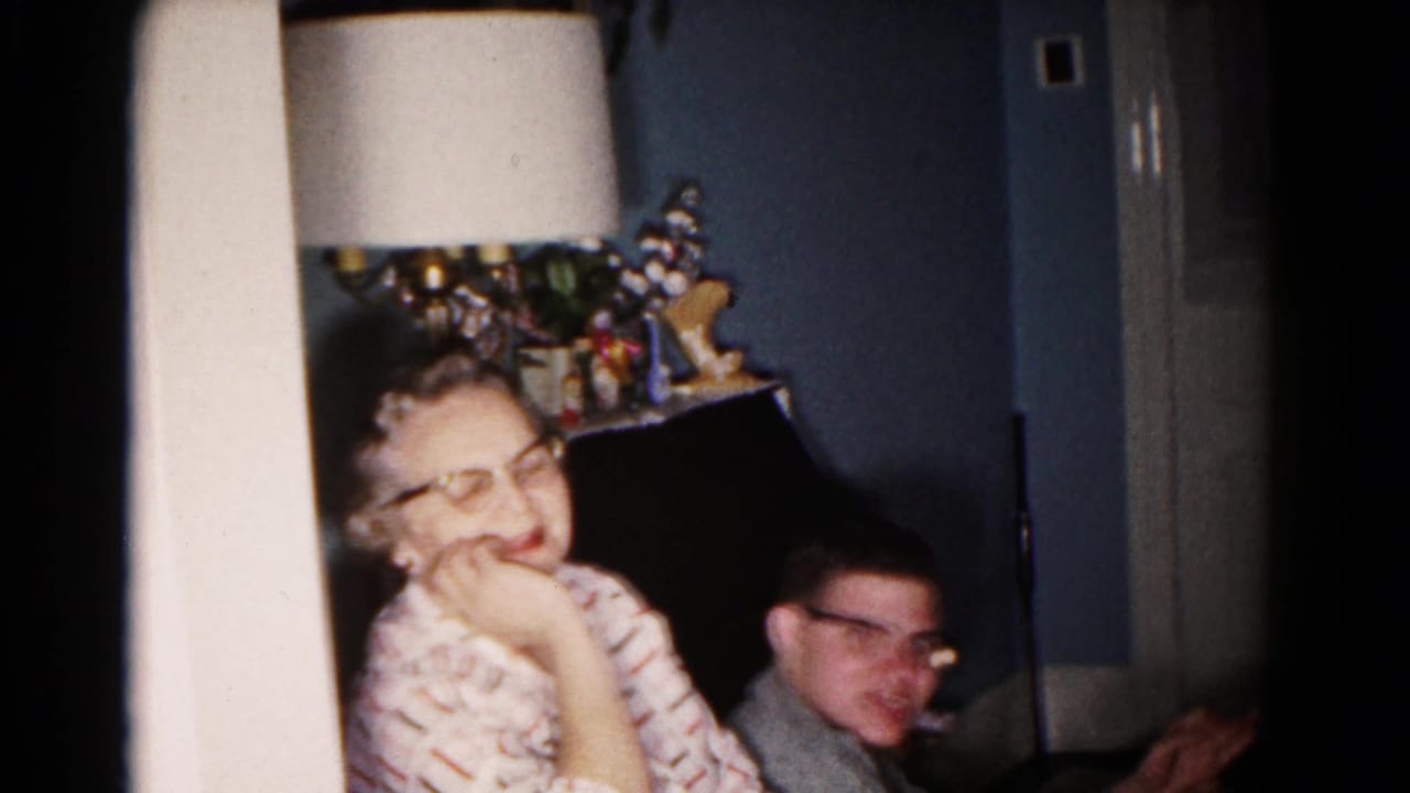 Inside the living room, a woman in patterned clothing and a man in a gray sweater sit near a tall lamp and dark piano-like furniture.