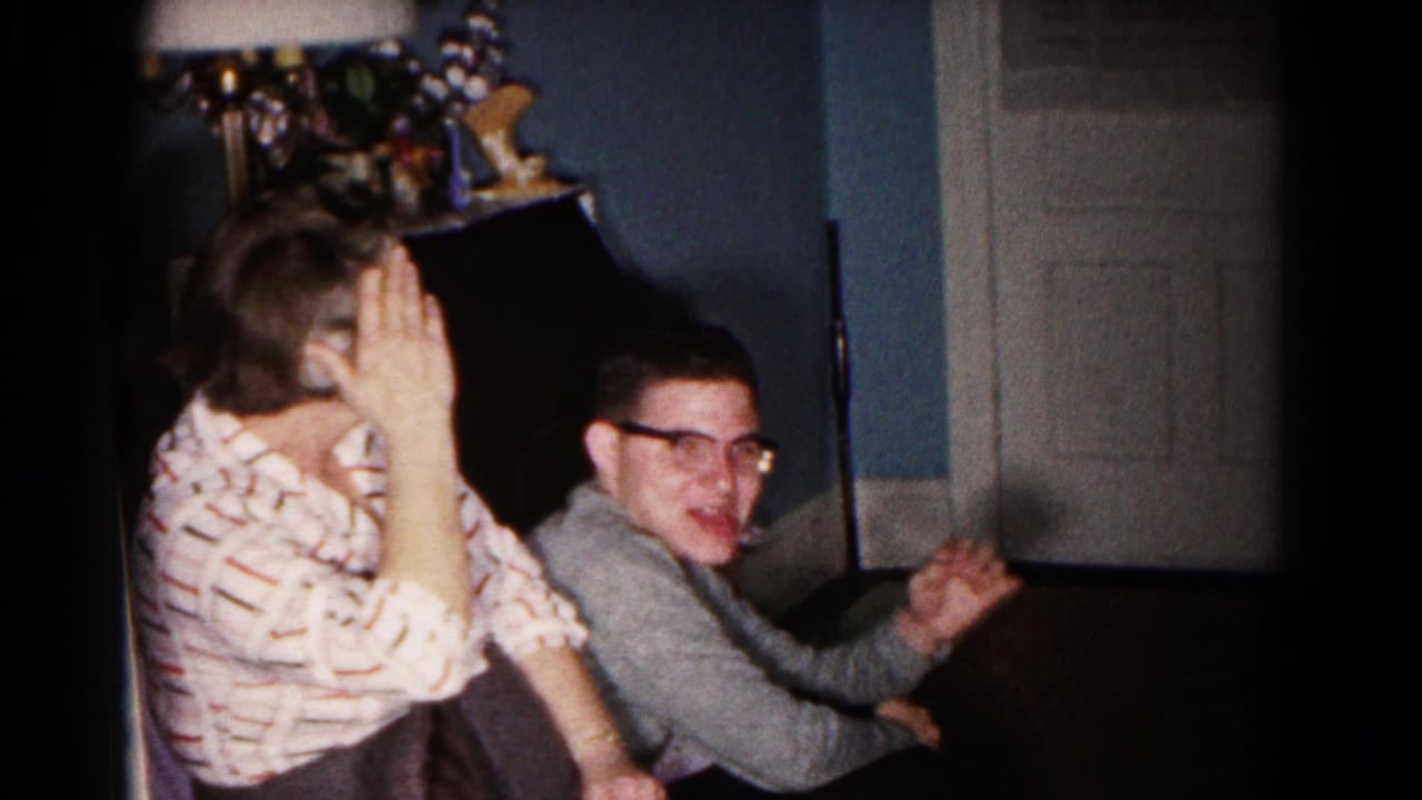 A seated woman raises a hand to her face as a smiling man in glasses looks toward the camera in the living room.