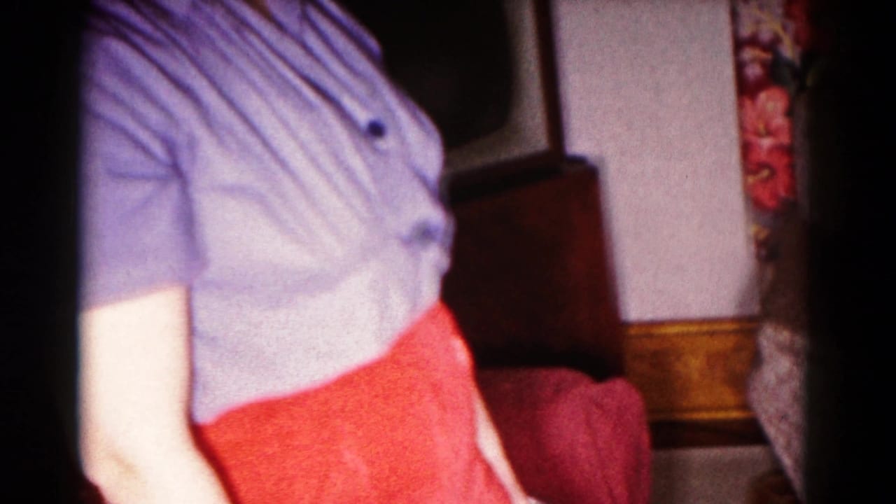 Person cropped at the torso standing indoors in a blue shirt and red skirt beside upholstered furniture.