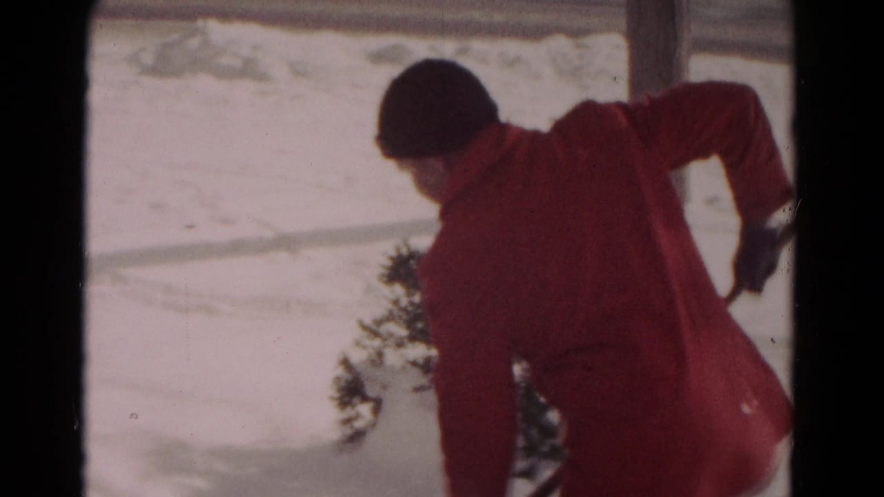 Person in a red coat bends over a snow shovel on a bright snowy surface.