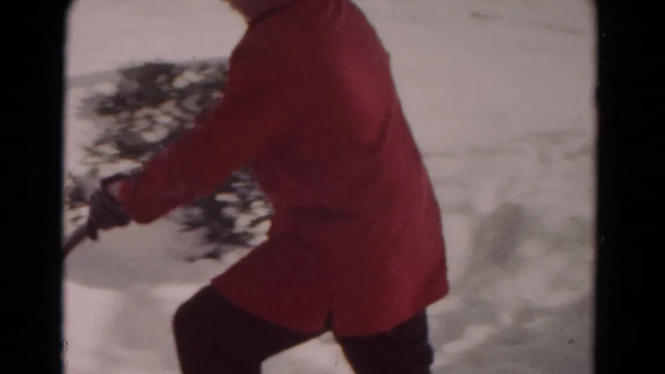 A side-angle 8mm frame shows the red-coated person moving a shovel through snow near low evergreen branches in Hagerstown, 1958.