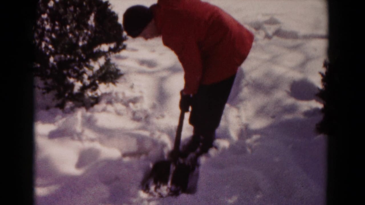 A 1958 Hagerstown winter yard scene shows a person leaning forward while shoveling snow near a bush.