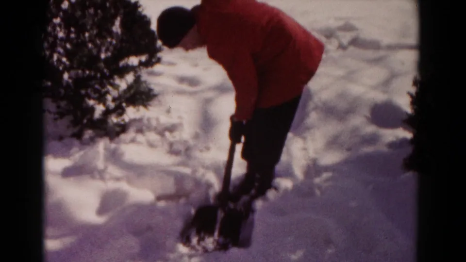 A 1958 Hagerstown winter yard scene shows a person leaning forward while shoveling snow near a bush.