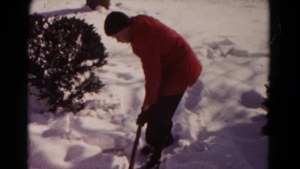 A person in an orange jacket shovels beside an evergreen bush in this 1958 Hagerstown winter home-movie frame.
