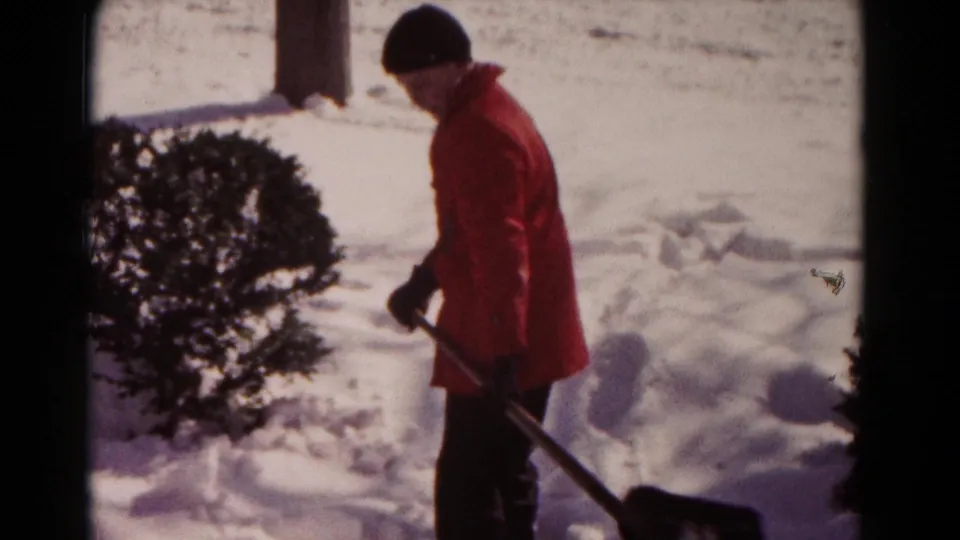 1958 Hagerstown footage shows a person standing with a shovel after working in deep snow near an evergreen bush.