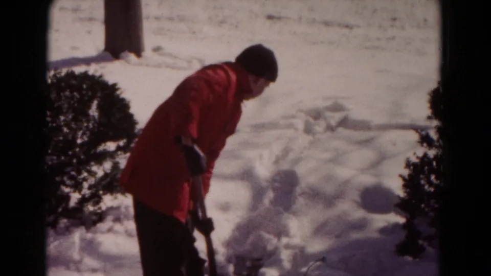 A red-coated person leans into the shoveling motion in snowy 1958 Hagerstown footage, partly framed by shrubs and winter glare.
