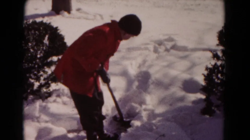 The person stands over the shovel near uneven snowbanks in the yard, captured in soft 8mm color with dark frame edges.