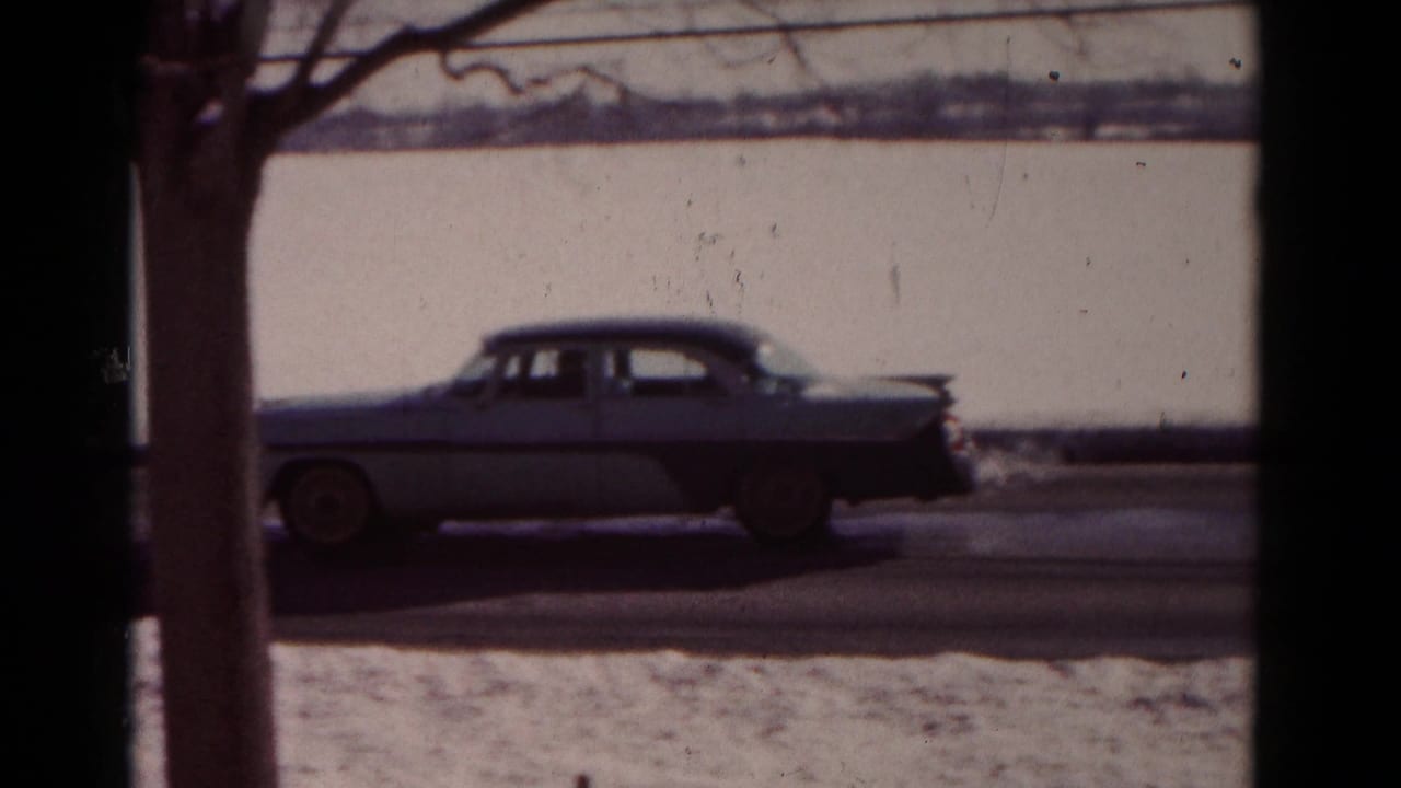 A two-tone 1950s sedan drives along a snow-covered street in Hagerstown, Maryland, seen through dark window framing.