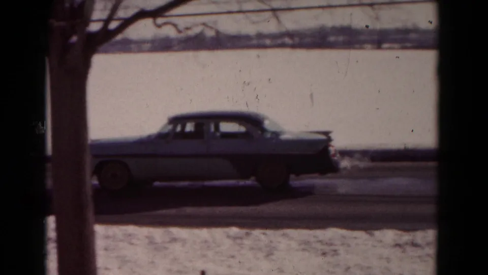 A two-tone 1950s sedan drives along a snow-covered street in Hagerstown, Maryland, seen through dark window framing.