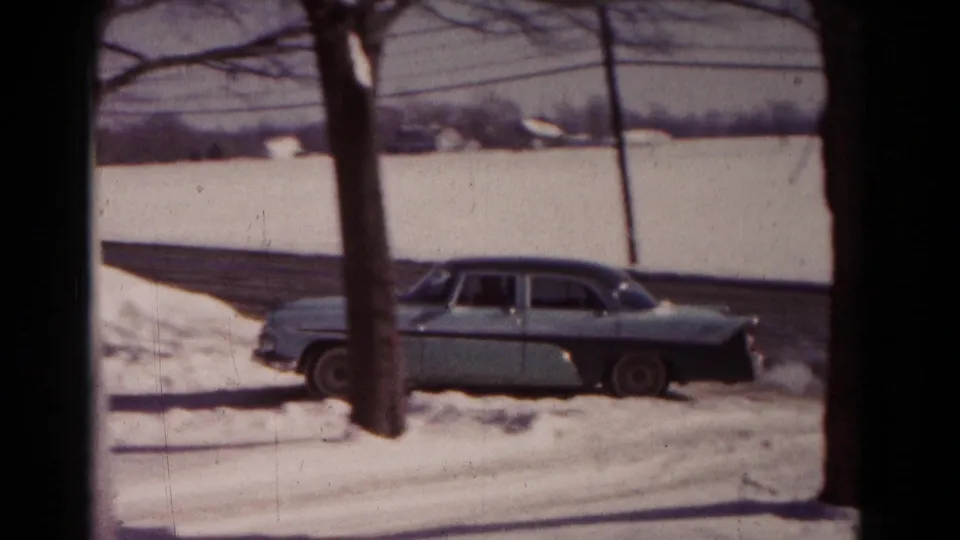 A side view of the 1950s sedan appears near a snowy curb and tree, with open winter landscape beyond.