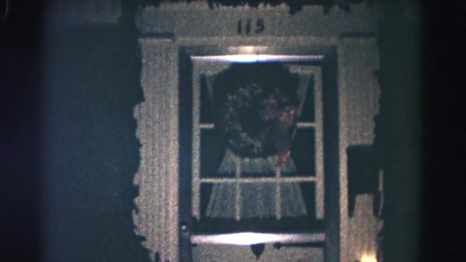 Close nighttime 1958 Hagerstown frame of a wreath in the front door window beneath house number 115, with curtains and a small light nearby.
