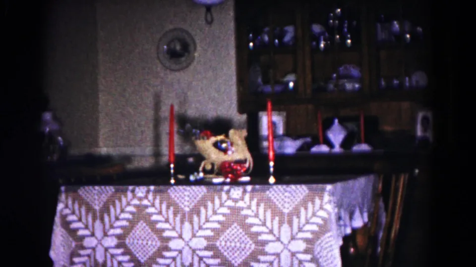 Low-light 8mm frame of a 1958 Hagerstown dining table with red candles, a centerpiece, patterned cloth, and cabinet in the background.