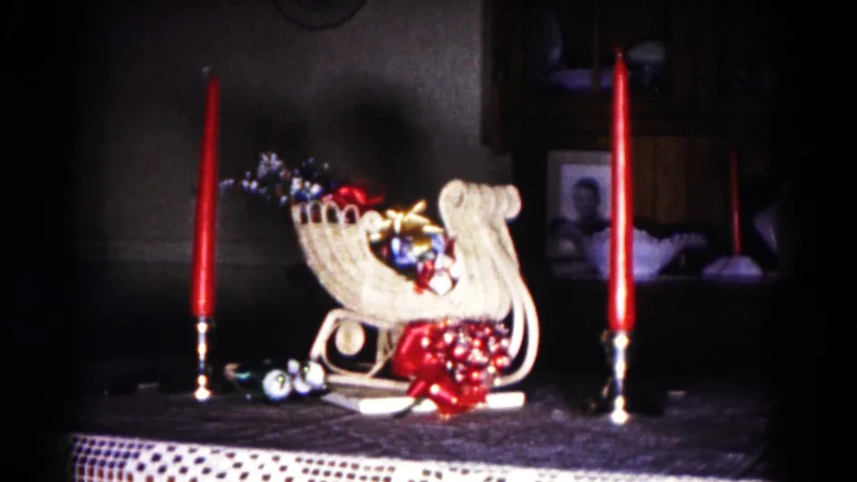 1958 Hagerstown frame of a white woven Christmas sleigh with ornaments and red bow, flanked by red candles on a tabletop.