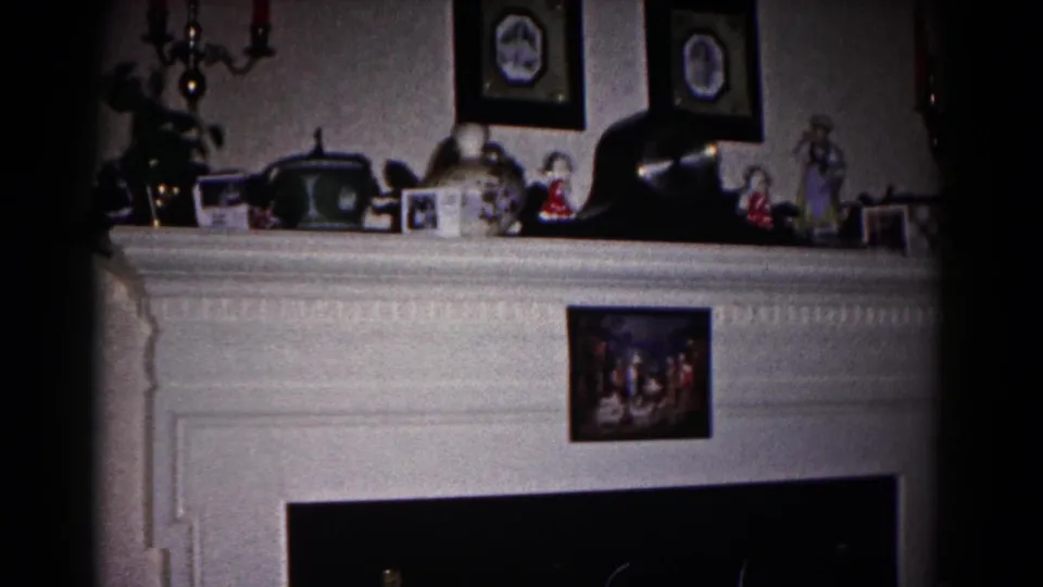 1958 Hagerstown home-movie frame of a white fireplace mantel with framed portraits, small ornaments, and a dark hearth below.