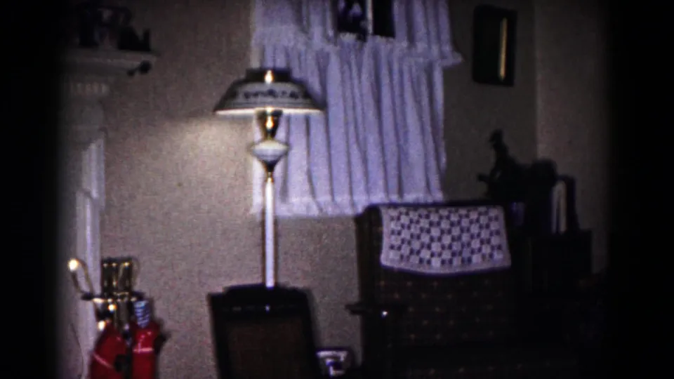 1958 Hagerstown home interior showing a lit floor lamp, curtained window, patterned chair, and crocheted throw.
