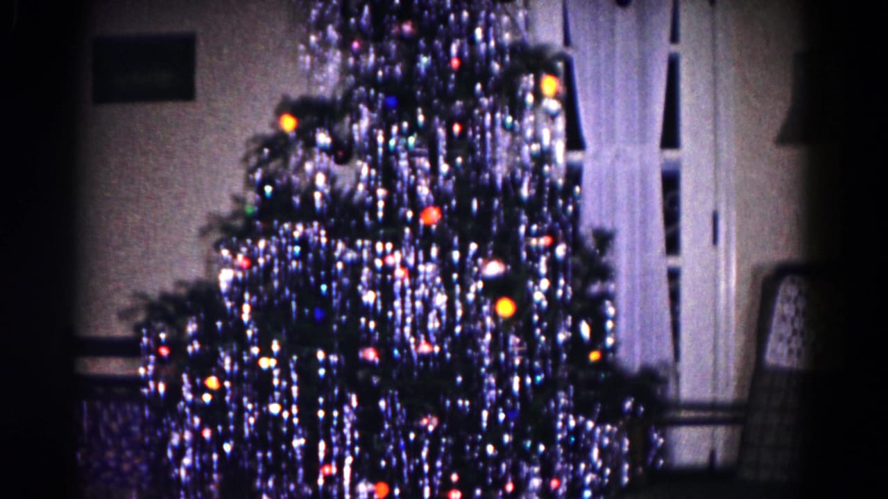 1958 Hagerstown interior frame with a tinsel-heavy Christmas tree, glowing colored bulbs, white curtains, and an upholstered chair.