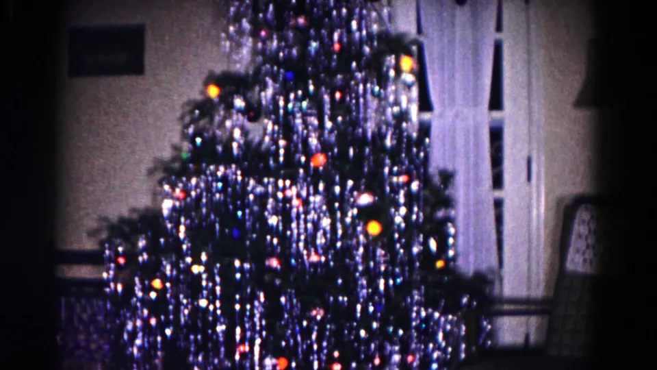 1958 Hagerstown interior frame with a tinsel-heavy Christmas tree, glowing colored bulbs, white curtains, and an upholstered chair.