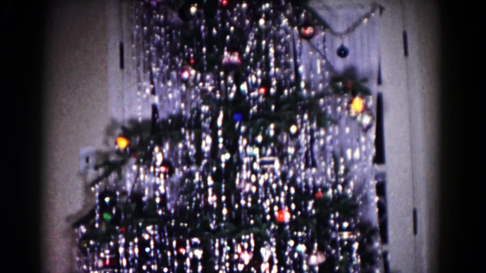 The Christmas tree remains framed indoors, showing dense silver tinsel, dark branches, colored bulbs, ornaments, and a door at the right.