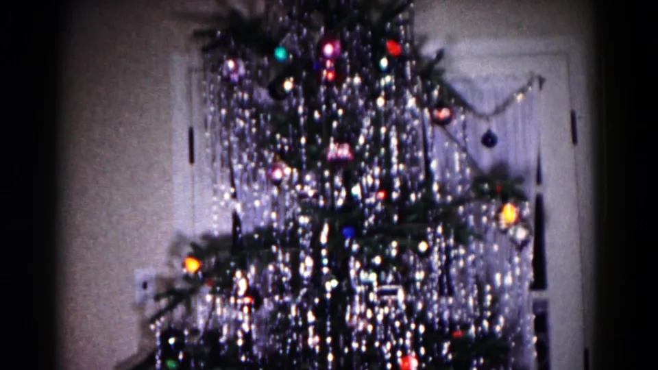 A soft 8mm frame shows the decorated indoor tree from a slightly lower angle, with sparkling tinsel and colored ornaments against the wall.