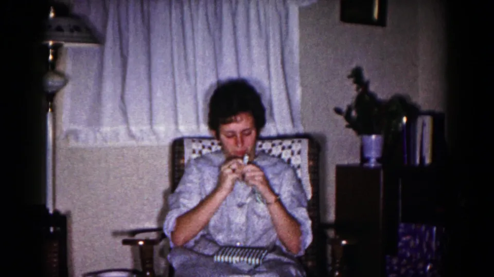 A woman in a light dress sits in a rocking chair in the Hagerstown living room, opening a small striped Christmas gift.