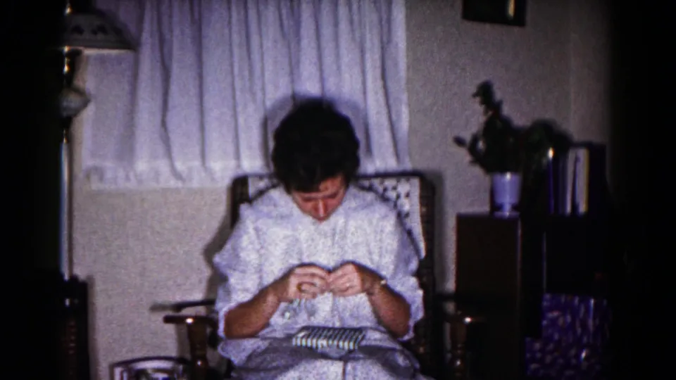The woman continues opening the striped present while seated in a rocking chair, with curtains, a lamp, and household decor behind her.