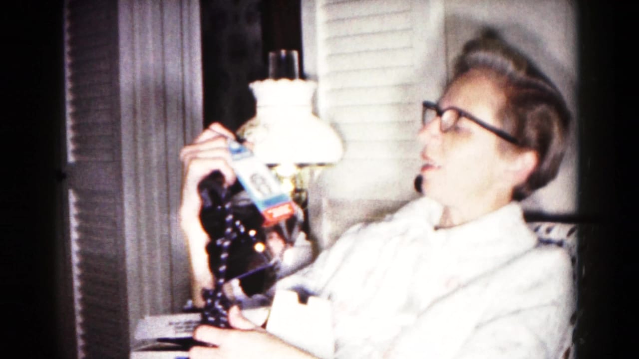 Woman in glasses seated indoors holding a dark electric iron near a lamp.