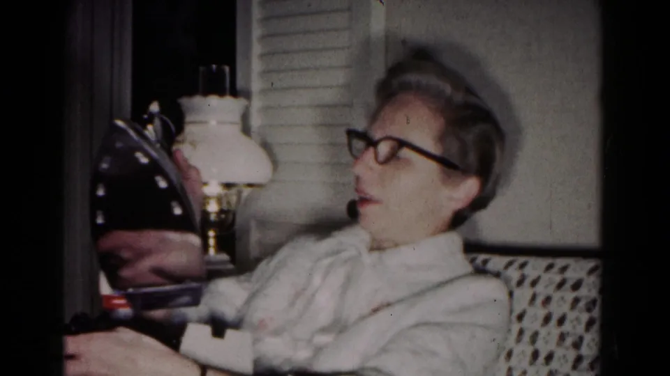 A woman in glasses sits on an upholstered sofa and inspects a dark iron in a softly exposed 1958 home-movie frame.