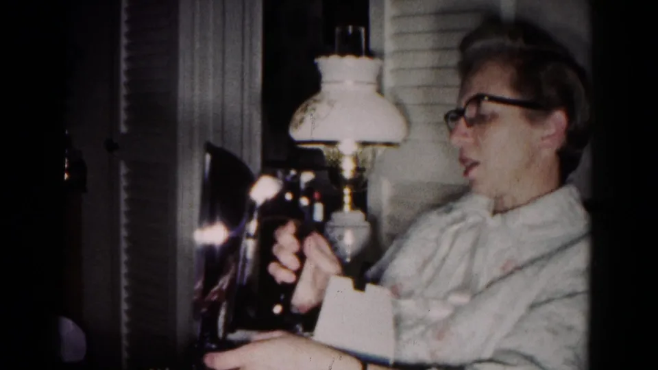 In a 1958 Hagerstown home interior, a woman in glasses holds a dark iron near a glowing lamp and curtains.