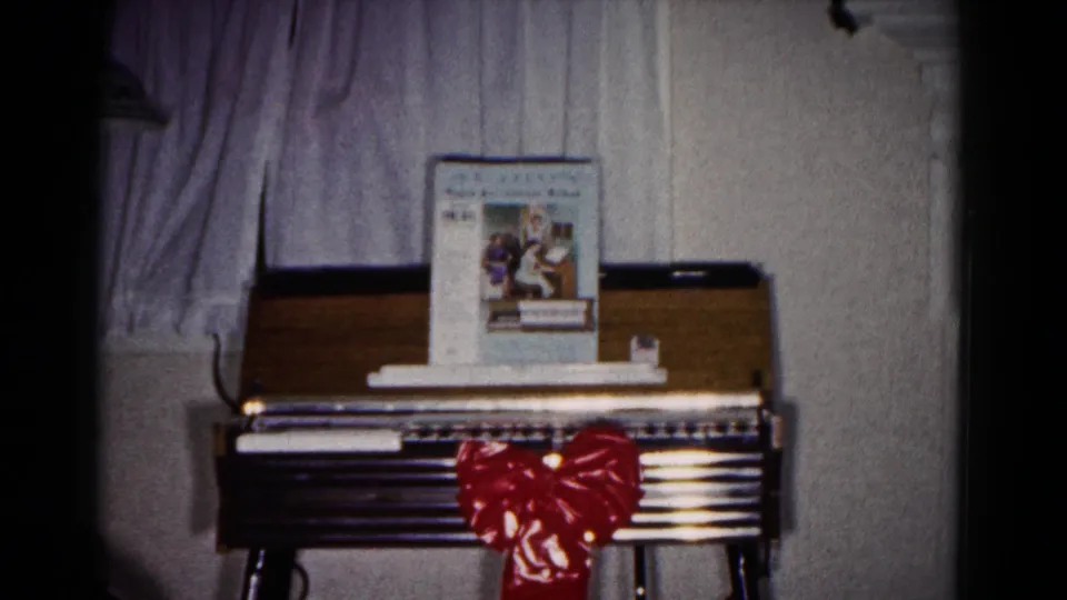A decorated keyboard instrument in a private home shows sheet music on top, curtains behind it, and a red bow across the front.