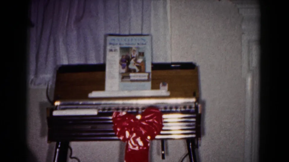 A soft 8mm frame shows the small keyboard instrument and red bow in the Hagerstown home, with the music book still visible above the keys.