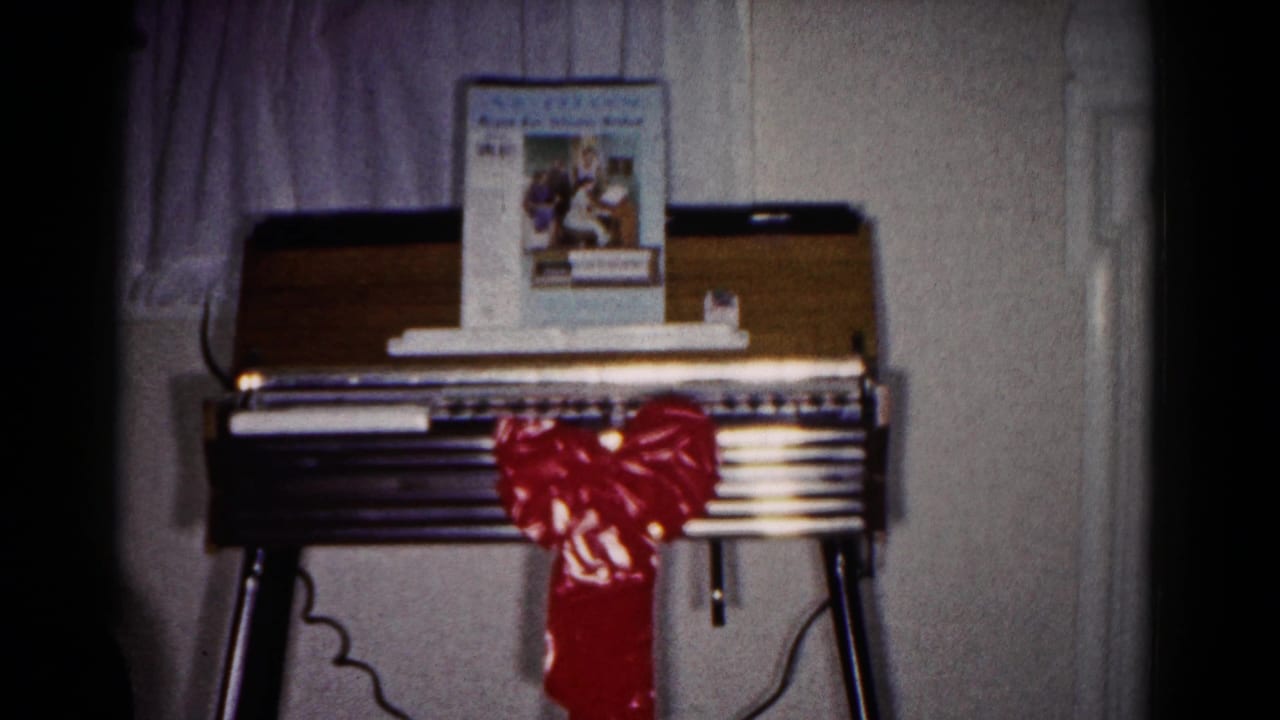 1958 Hagerstown home-movie frame showing a wooden organ with a red Christmas bow, music book, curtains, and wall paneling.