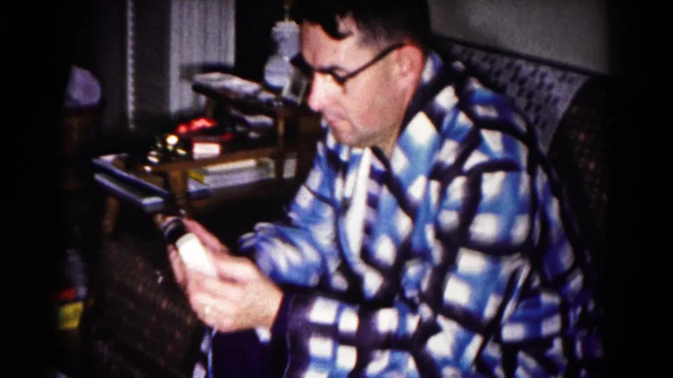 1958 Hagerstown home-movie frame of a seated man in glasses and a blue plaid robe focused on a small object in his hands.