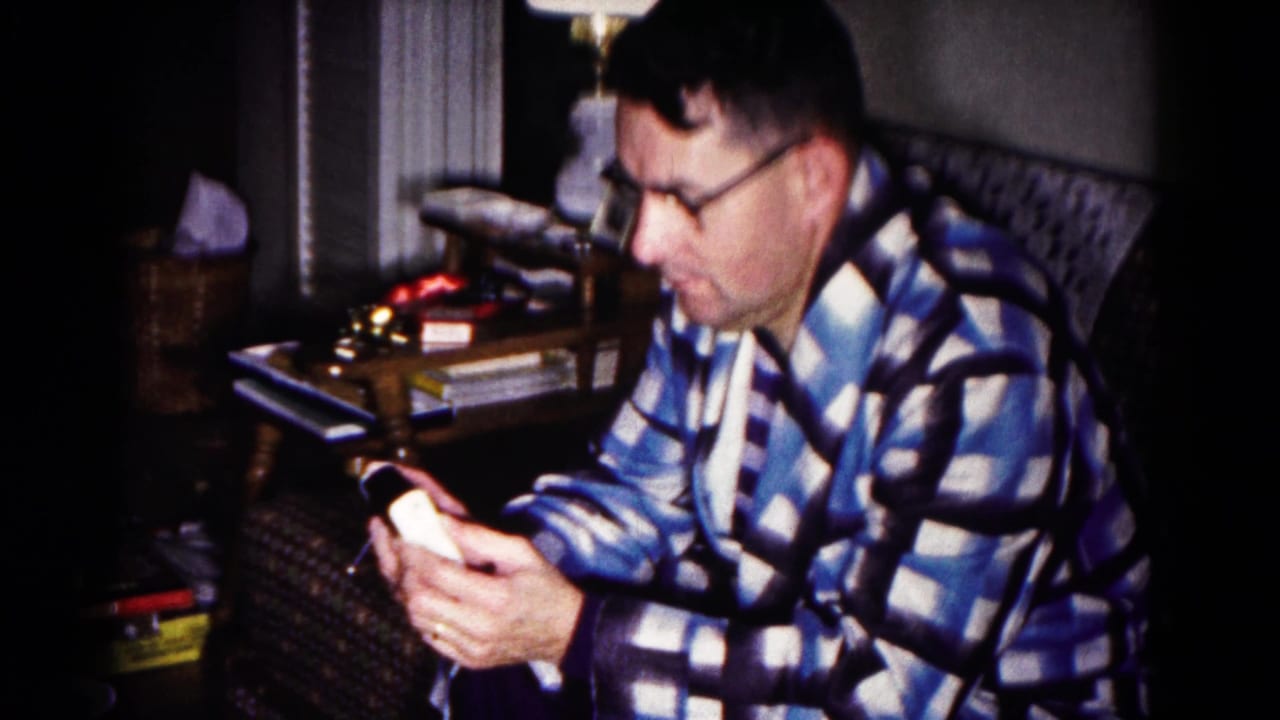 Man in glasses and a check robe looks down at a small device in a dim living room.
