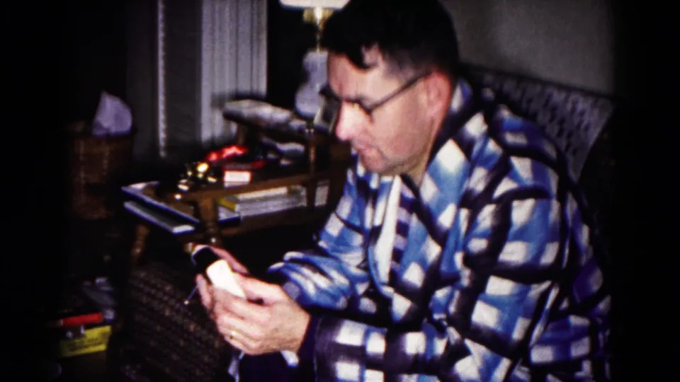 In 1958 Hagerstown home-movie footage, a man in glasses and a blue check robe studies a small handheld device beside a lamp and side table.