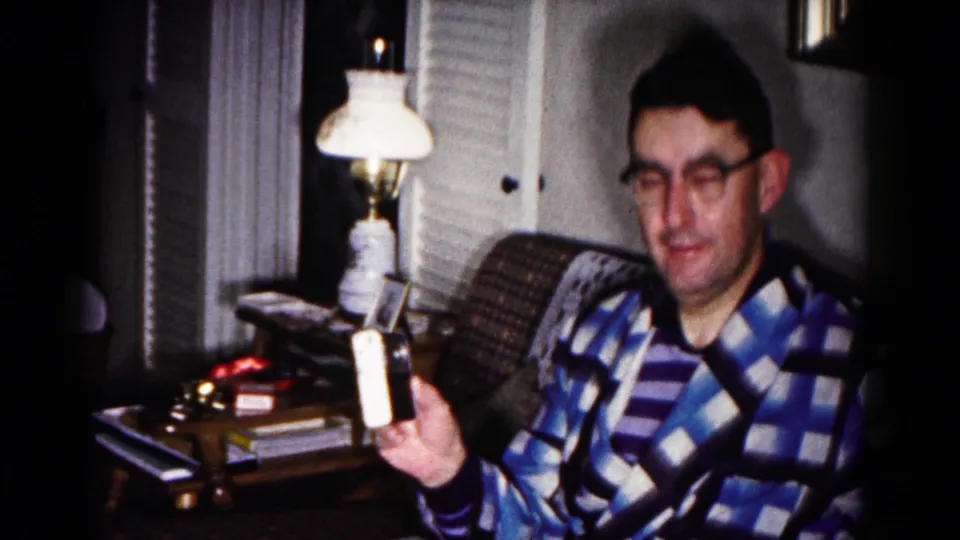 The man in glasses holds the small device outward toward the camera, with a lamp, chair, and side table visible in the 1958 interior.