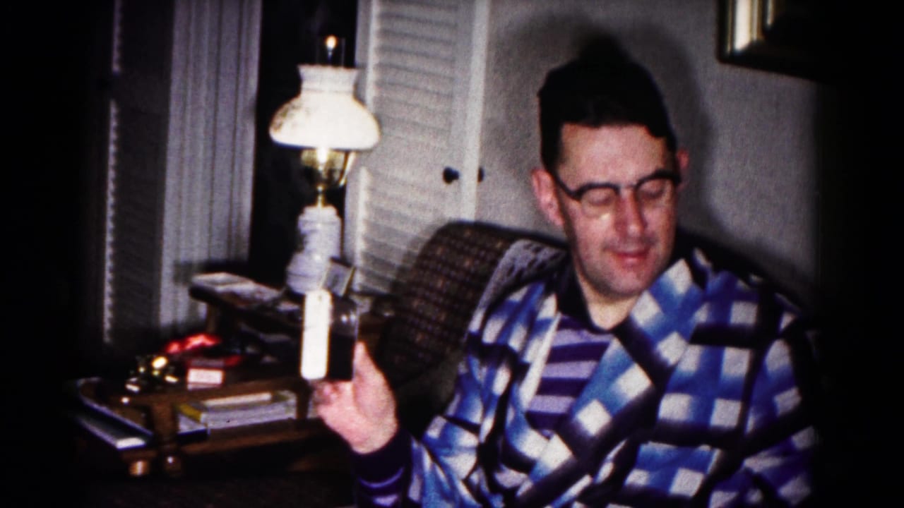 In a 1958 Hagerstown home interior, a seated man in glasses and a blue checkered robe holds up a small white object near a table lamp.