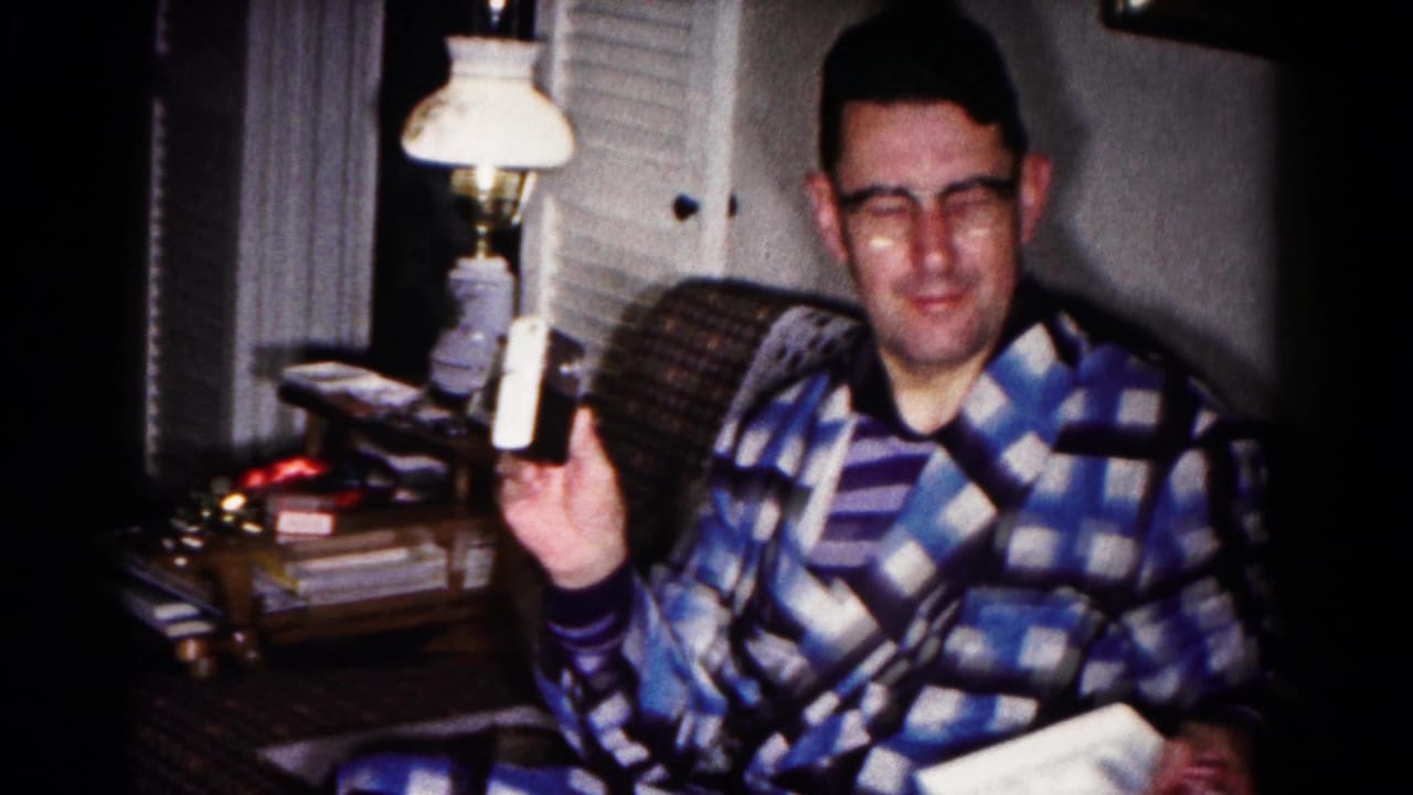 The man in glasses smiles in a dim Hagerstown living room while lifting a white cylindrical object and holding an open gift box.