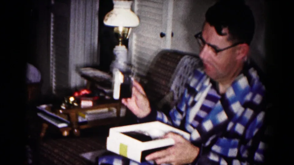 Inside the 1958 home setting, the man turns toward the white object while an open box rests in his hands near a lamp and side table.