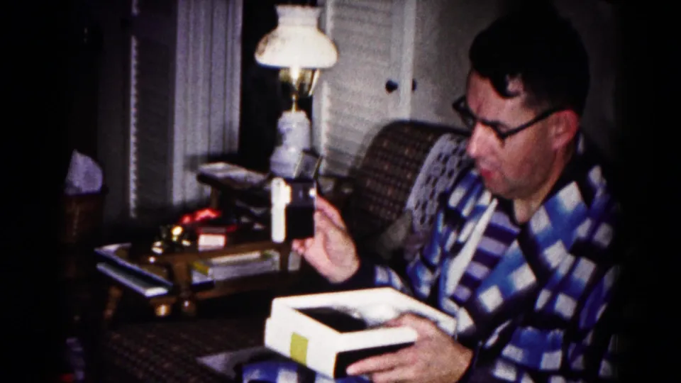 The seated man in the Hagerstown home-movie frame studies the white object while holding an open box under warm lamp light.
