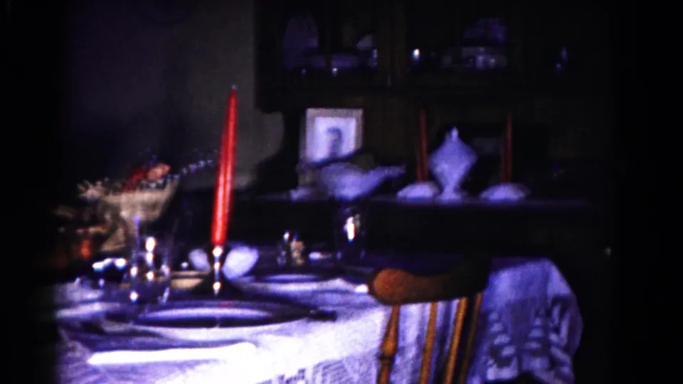 1958 Hagerstown home-movie frame showing a dim formal dining table with red candles, table settings, and background shelving.