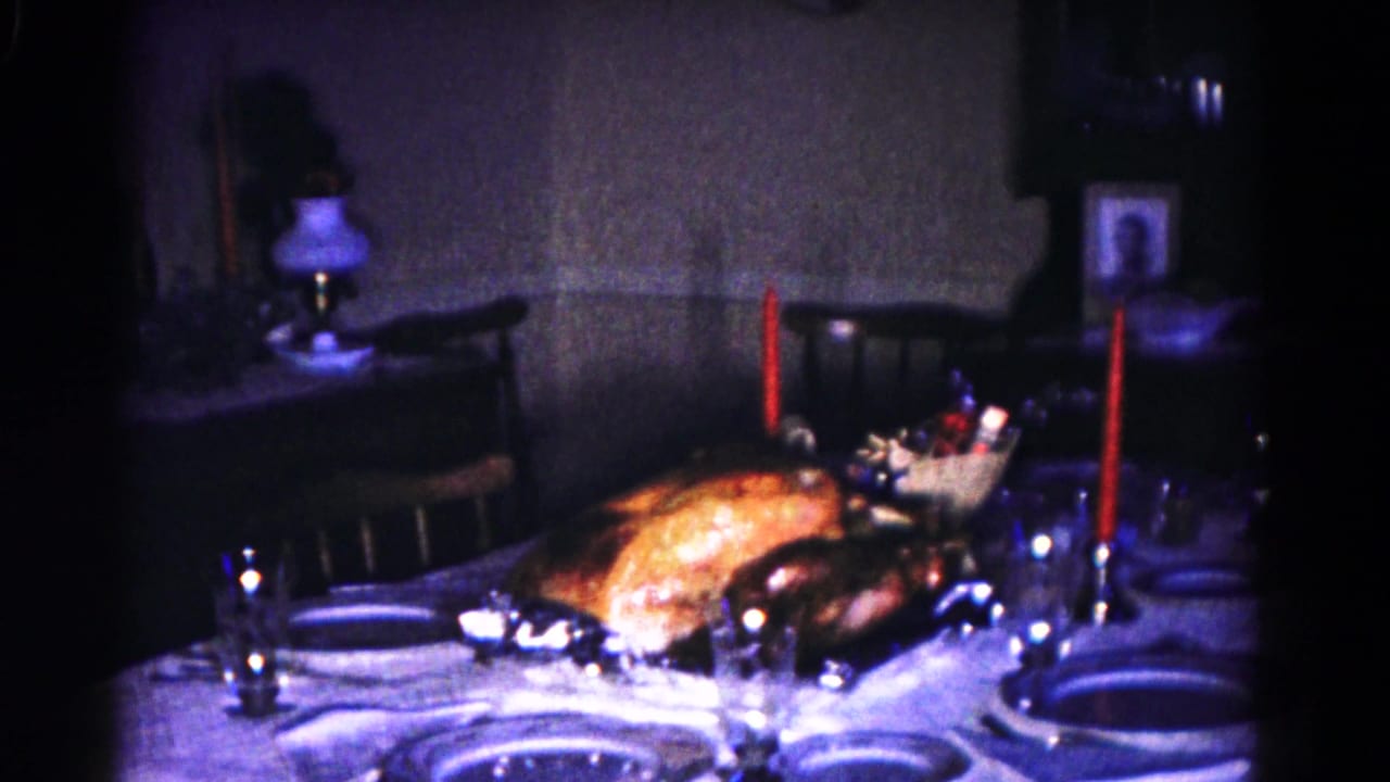 Dim dining table with roast turkey, plates, glasses, red candles, lamp, and framed portrait.