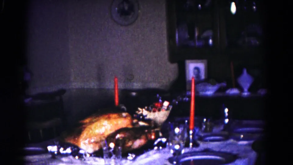 1958 8mm frame of a private dining room with the turkey and red candles nearer the foreground, surrounded by plates, glasses, and utensils.
