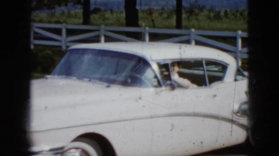 1958 Las Vegas home-movie frame showing a white vintage sedan by a white fence, with a woman partly visible inside.
