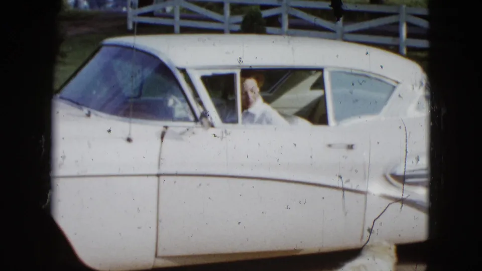 1958 Las Vegas home-movie still showing a woman in a white blouse visible in a white sedan beside a white fence.